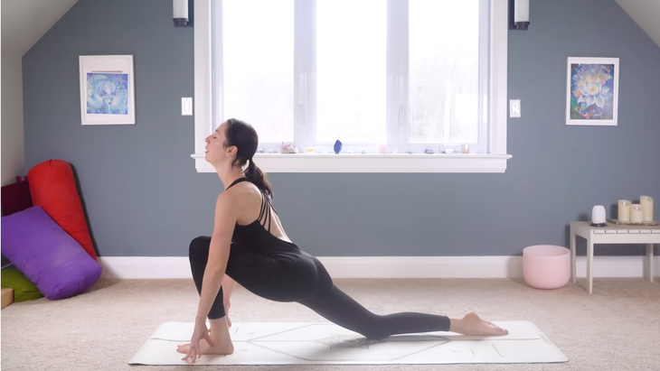 woman in black yoga clothes in Low Lunge Pose with left foot forward on tan yoga mat on carpeted floor of home with window and blue wall behind her