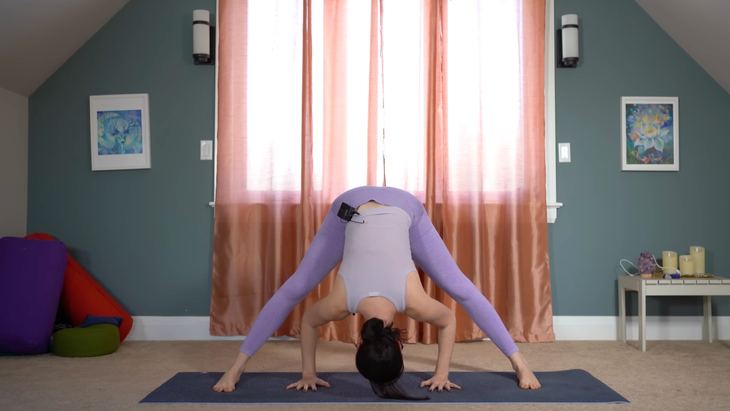 woman in purple yoga clothes in wide-legged standing forward bend facing the side of her mat with hands underneath her and top of head resting on mat