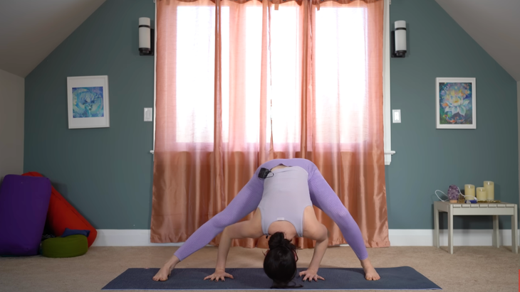 woman in purple yoga pants in wide-legged standing forward fold with head and hands on ground below her bends left knee