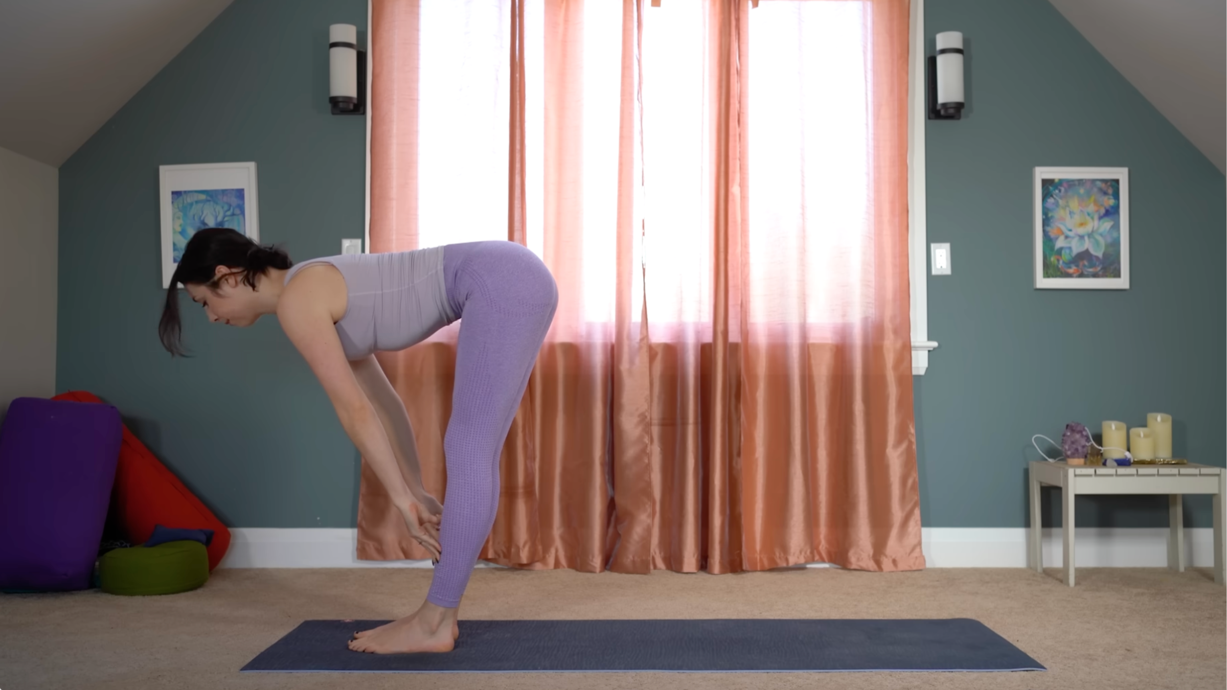 woman in purple yoga clothes stands in half forward bend pose with fingertips on shins at front of mat