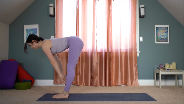 woman in purple yoga clothes stands in half forward bend pose with fingertips on shins at front of mat
