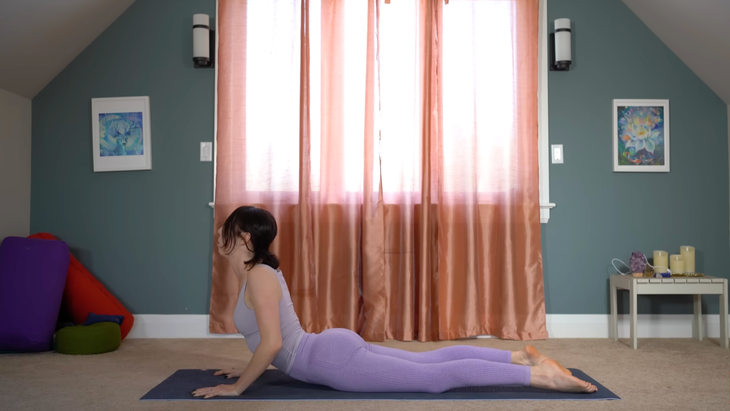 woman in purple yoga clothes in cobra pose on yoga mat in house in front of window with closed curtains