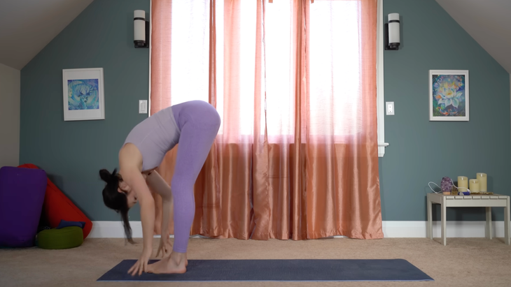 woman in purple yoga clothes in standing forward fold at front of yoga mat in house in front of window with closed curtains