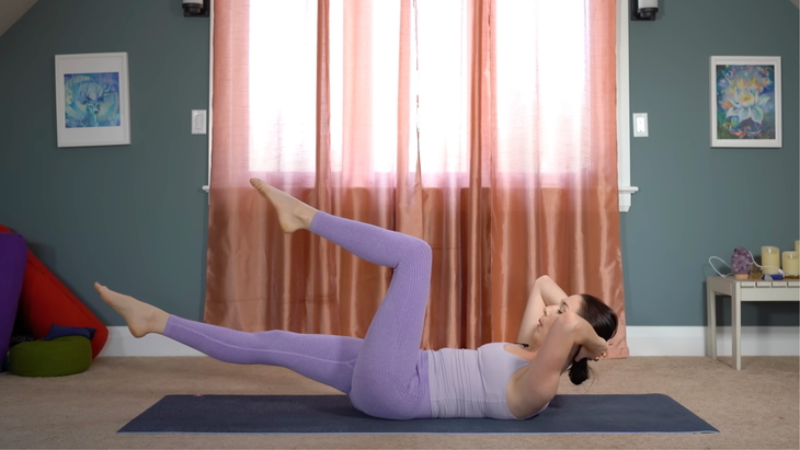 woman in purple yoga clothes in half boat variation with head and shoulders raised and hands clasped behind head, right leg extended and hovering above ground and left knee bent