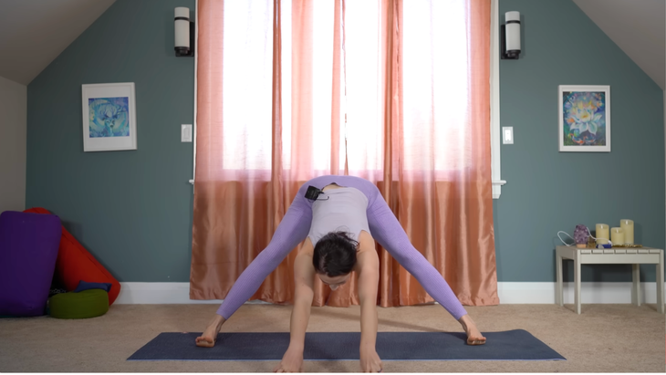 woman in purple yoga clothes in wide-legged standing forward fold facing the side of her mat with arms on ground in front of her