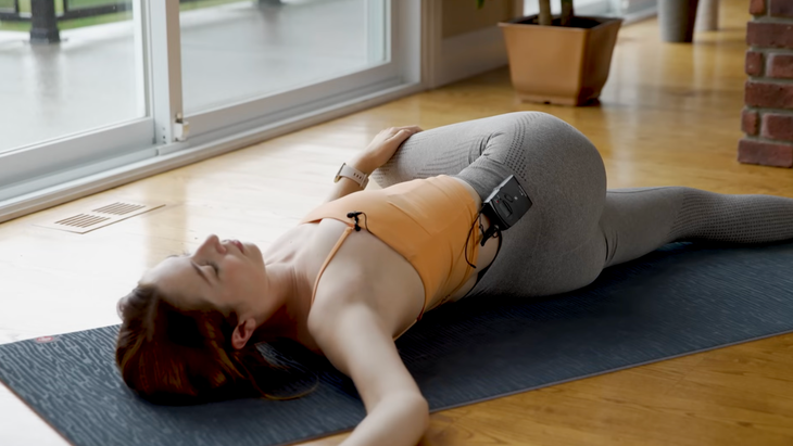 woman in orange tank top and gray leggings in reclined twist pose lying down on gray yoga mat right leg crossed over to left side of mat, left hand on right knee, right arm bent and relaxed on right side of mat, face toward ceiling, on wood floor in home with brick wall, sliding glass doors, and plants in the background