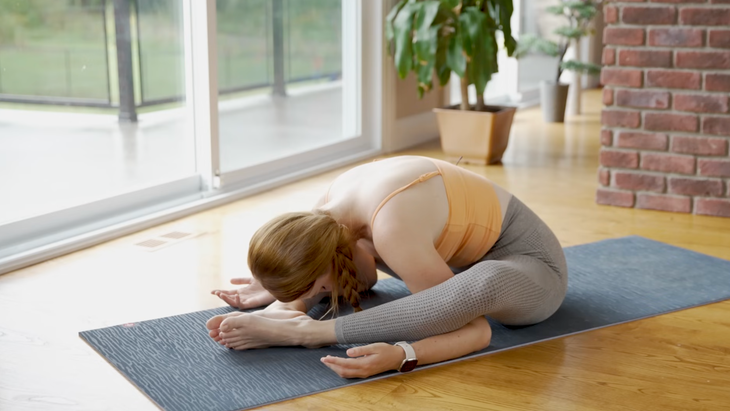 woman in orange tank top and gray leggings sits in turtle pose, seated on gray yoga mat leaning head toward mat with arms under bent knees, on wooden floor in home with sliding doors and plants in background
