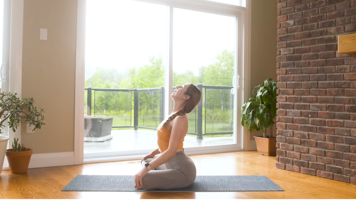 woman in orange tank top and gray leggings sits in seated cat-cow pose, sitting cross-legged on gray yoga mat with hands on knees and face and chest toward the ceiling, on wooden floor in home with sliding doors and plants in background