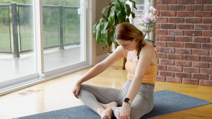 woman in orange tank top and gray leggings sits in seated cat pose, sitting cross-legged on gray yoga mat with hands on knees, back curved and face looking down toward angles, on wooden floor in home with sliding doors and plants in background