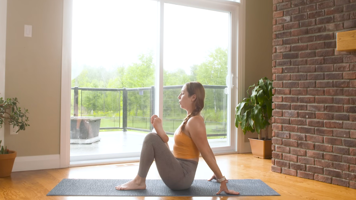 woman in orange tank top and gray leggings sits in seated pigeon pose, sitting on gray yoga mat leaning back on hands, left foot on floor, and right ankle crossed over right thigh, on wooden floor in home with sliding doors and plants in background
