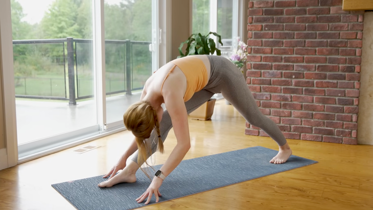 woman in orange tank top and gray leggings in pyramid pose varation with feet spread wide forward and backward on gray yoga mat, legs straight, and body folded forward over right leg with fingertips touching the mat, on wood floor in home with brick wall, sliding glass doors, and plants in the background