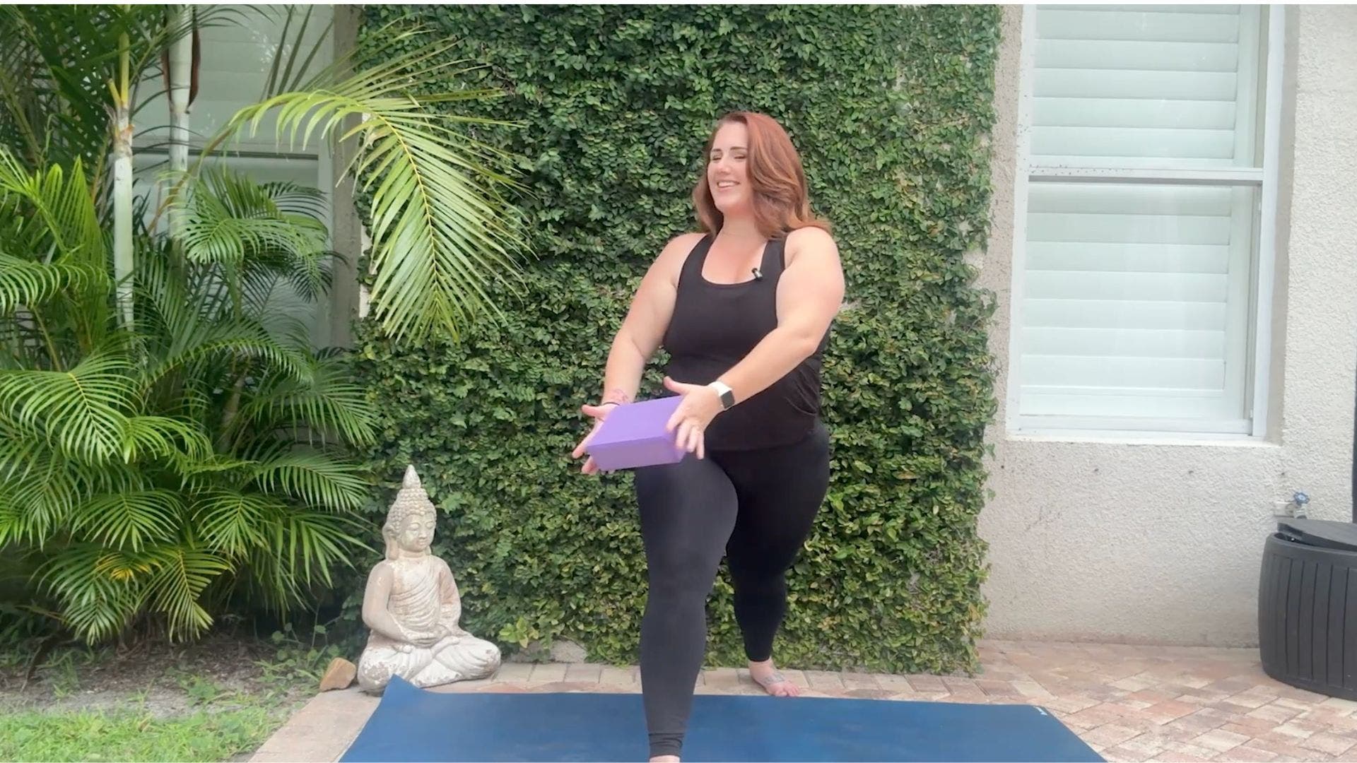 Woman on yoga mat in Warrior 1 holding purple yoga block between her hands.