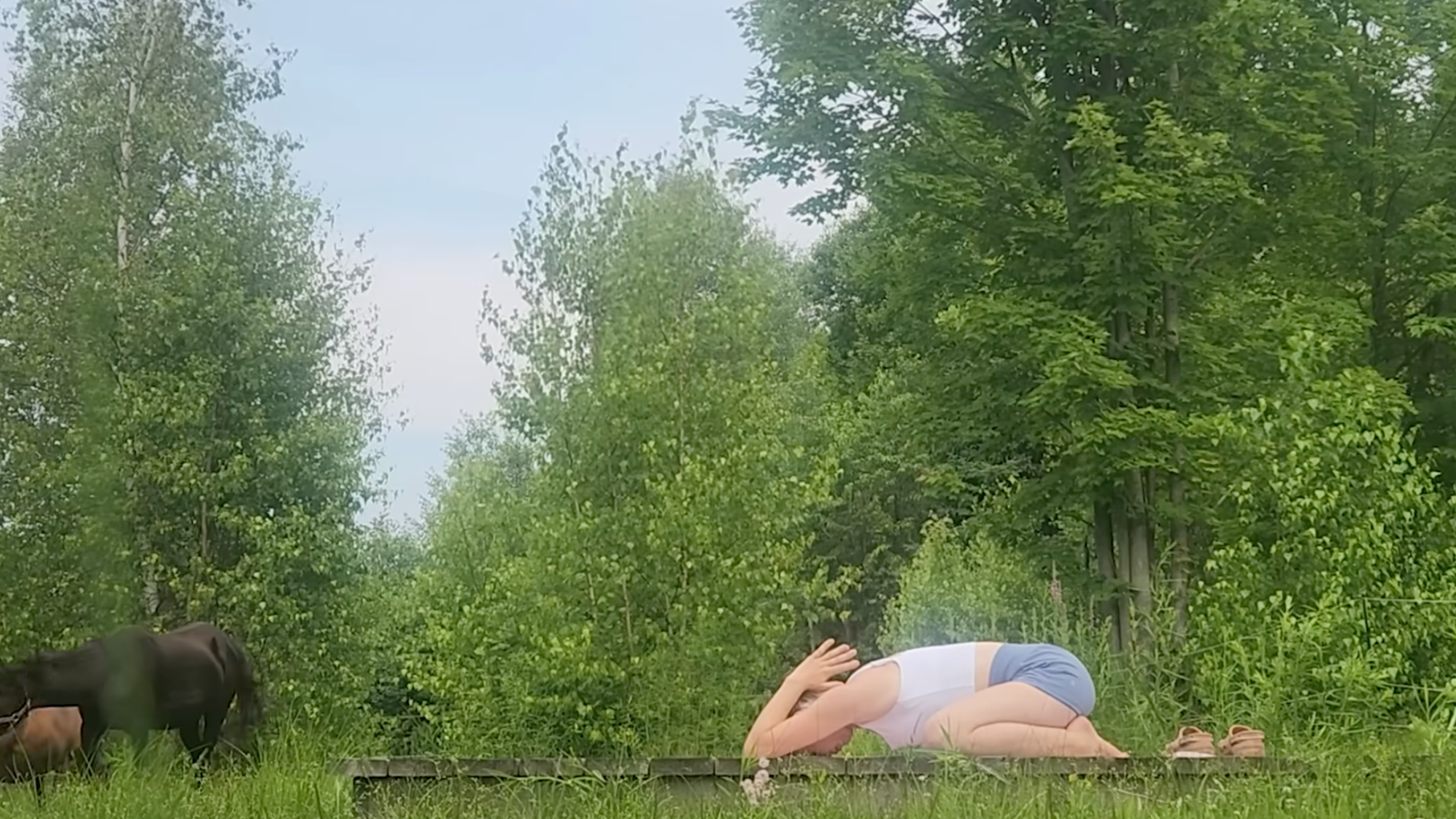 woman in child pose on wooden platform with trees and horses in the background