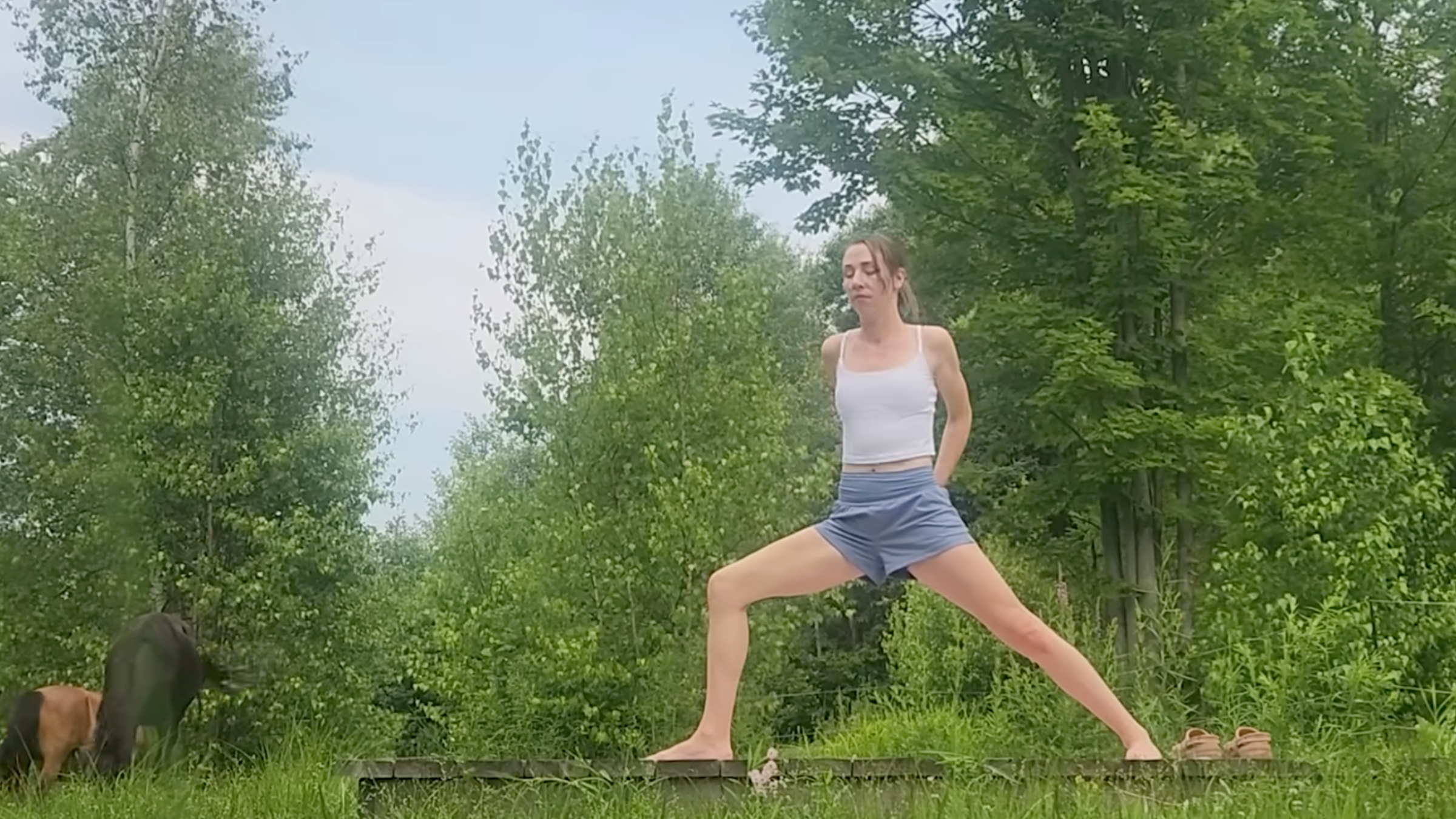 woman prepares for humble warrior yoga pose by interlacing fingers behind back outside on wooden platform in front of trees and horses