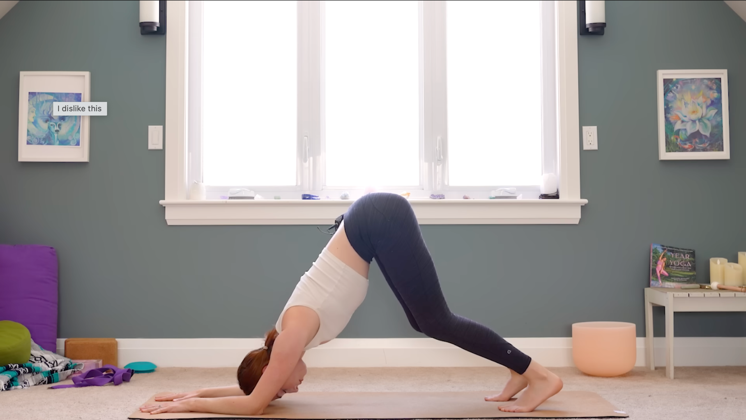 woman in white tank and black tights in dolphin pose with forearms on ground, forehead on mat, and hips raised toward the ceiling, on tan yoga mat in carpeted room with windows and yoga accessories in the background