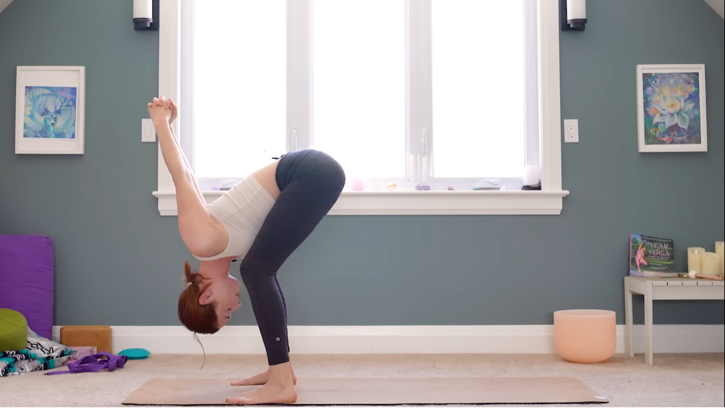 woman in white tank and black tights in forward fold with knees bent generously and fingers interlaced behind back and lifted toward the ceiling, on tan yoga mat in carpeted room with windows and yoga accessories in the background