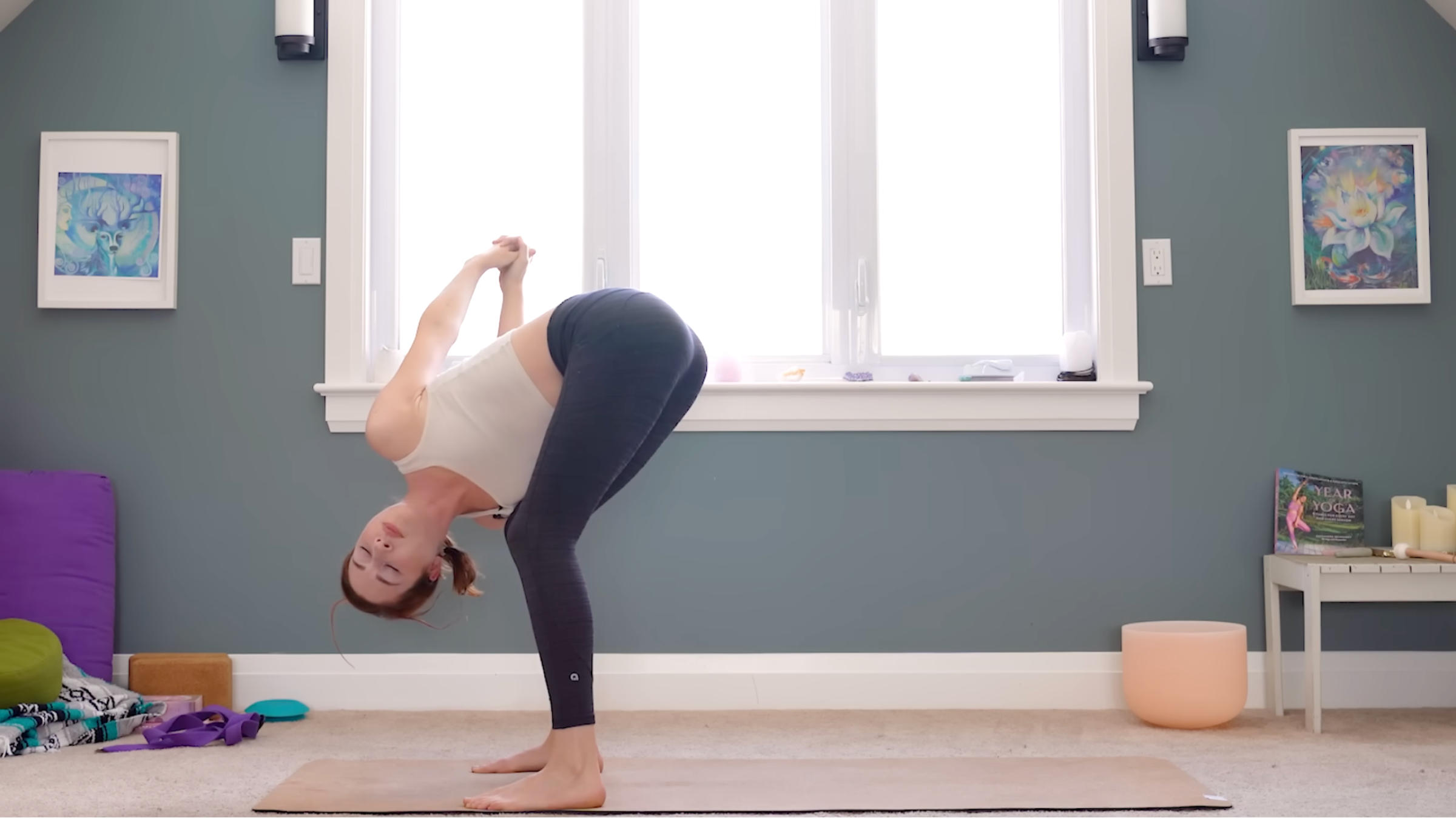woman in white tank and black tights in standing forward bend pose with knees bent, arms interlaced behind back, and right shoulder on right thigh with spine twisted, on tan yoga mat in carpeted room with windows and yoga accessories in the background