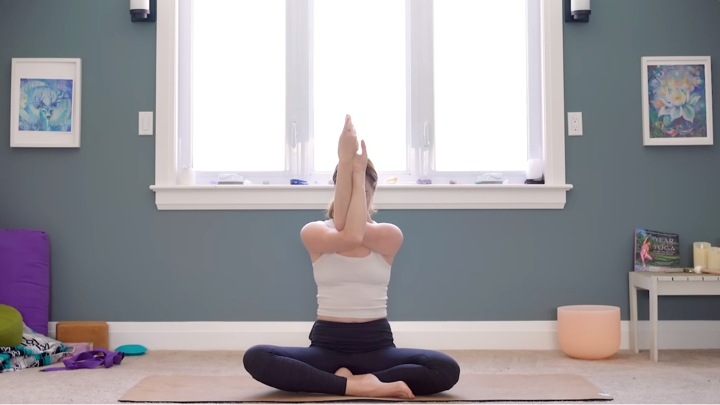 woman in white tank and black tights seated cross-legged seat with arms in eagle pose as shoulder stretch, on tan yoga mat in carpeted room with windows and yoga accessories in the background