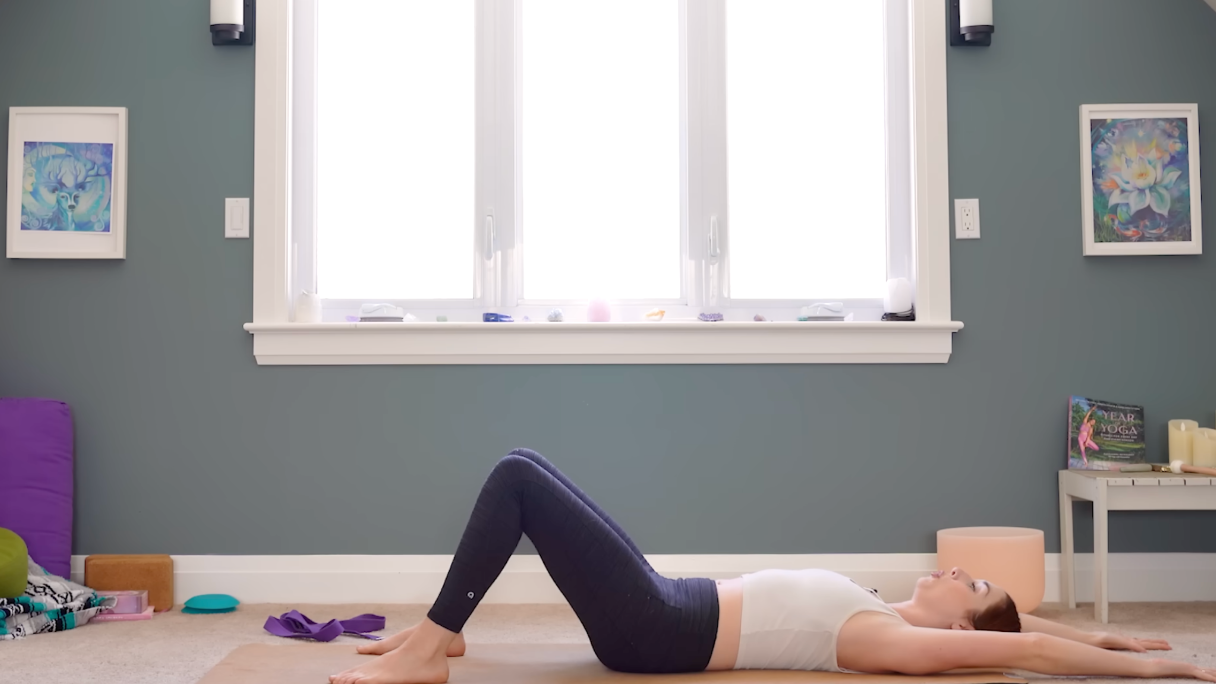 woman in white tank and black tights in reclined shoulder stretch pose with knees bent, feet on floor, arms stretched on floor past head, on tan yoga mat in carpeted room with windows and yoga accessories in the background