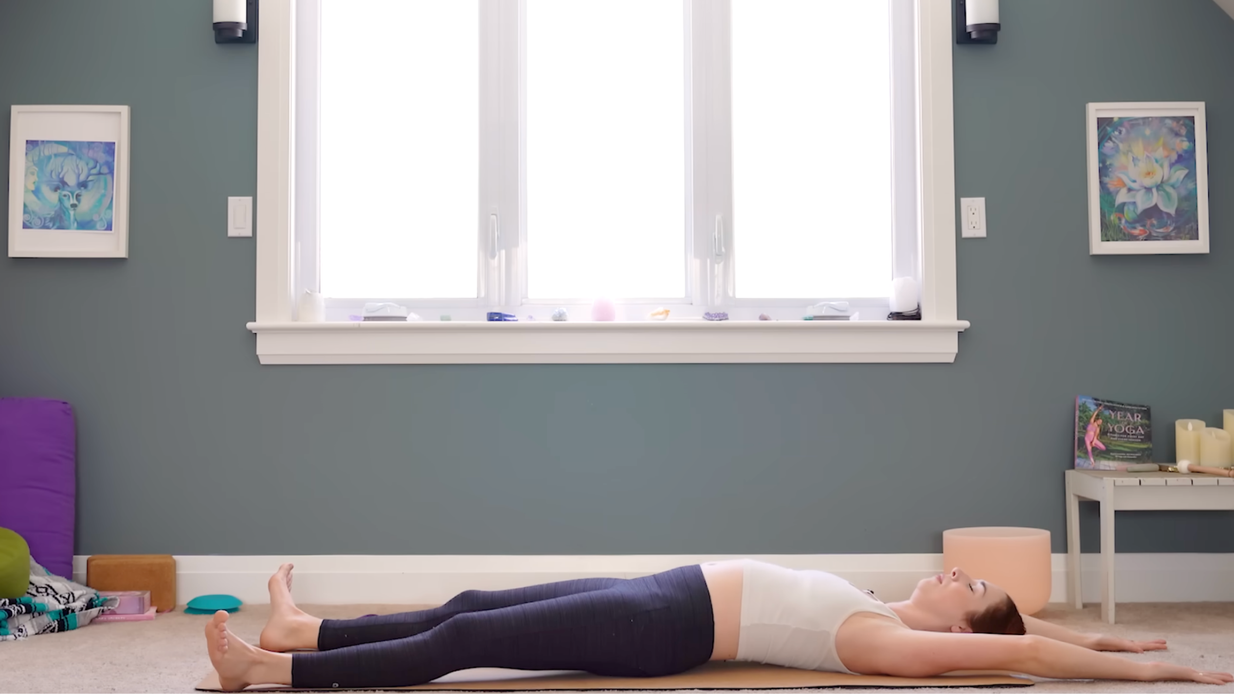 woman in white tank and black tights in reclined shoulder stretch pose with legs outstretched, arms stretched on floor past head, on tan yoga mat in carpeted room with windows and yoga accessories in the background
