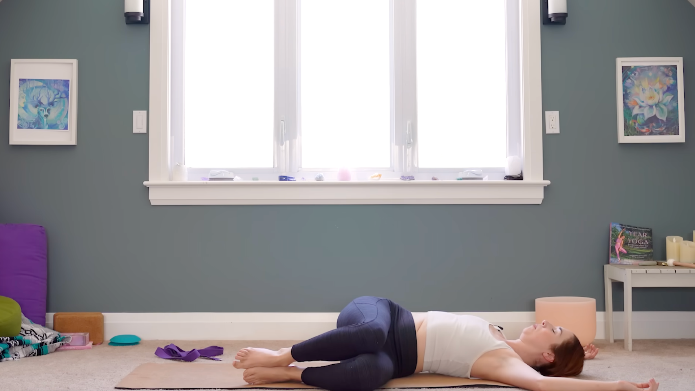 woman in white tank and black leggings lying in reclined spinal twist pose with knees bent and legs to the left and cactus arms on the ground on a tan yoga mat in room with windows and yoga accessories in the background
