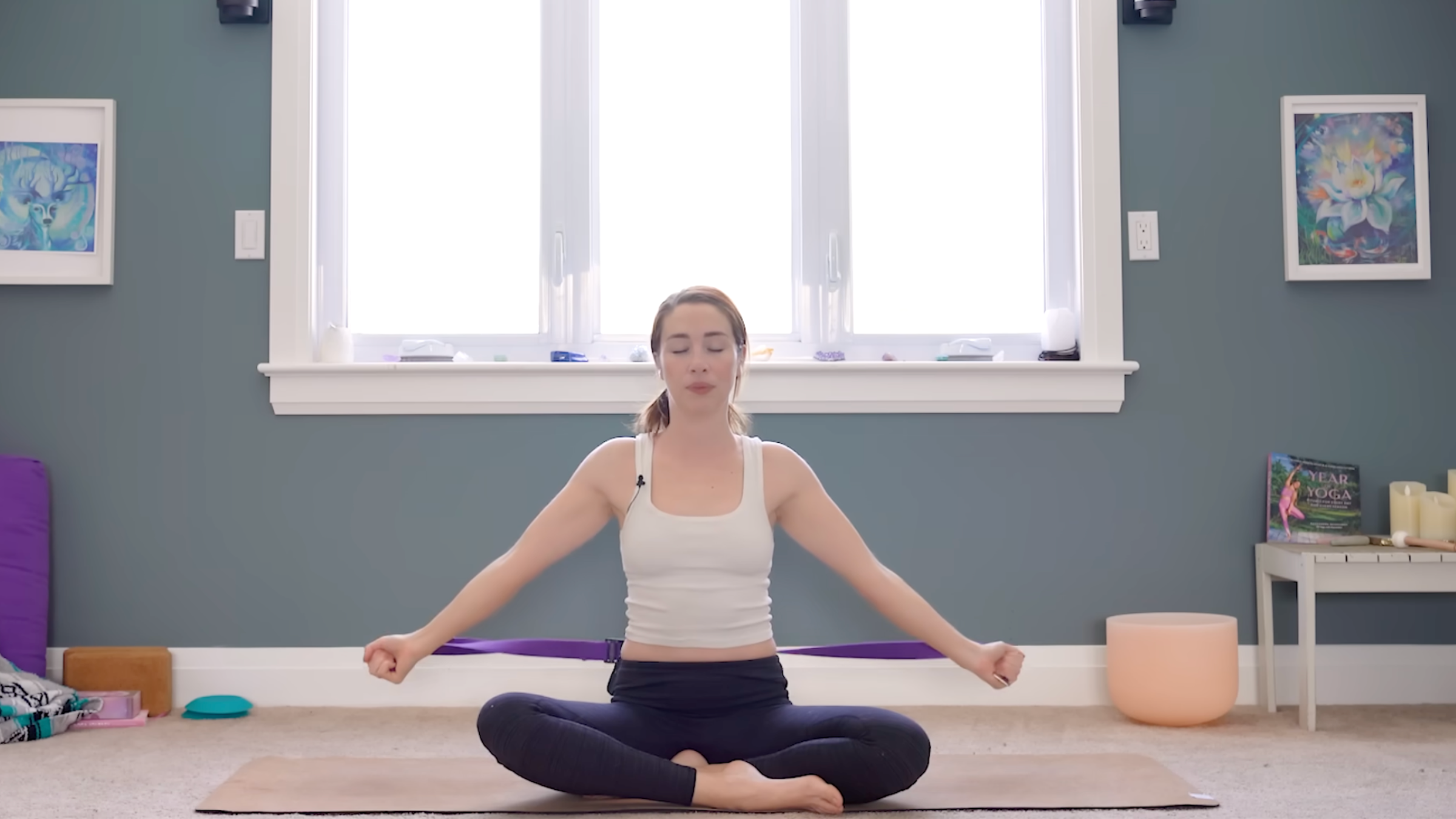 woman in white tank and black leggings sits cross-legged on yoga mat with hands grabbing the ends of purple band, arms straight and lowered by hips, in room with windows and yoga accessories in the background