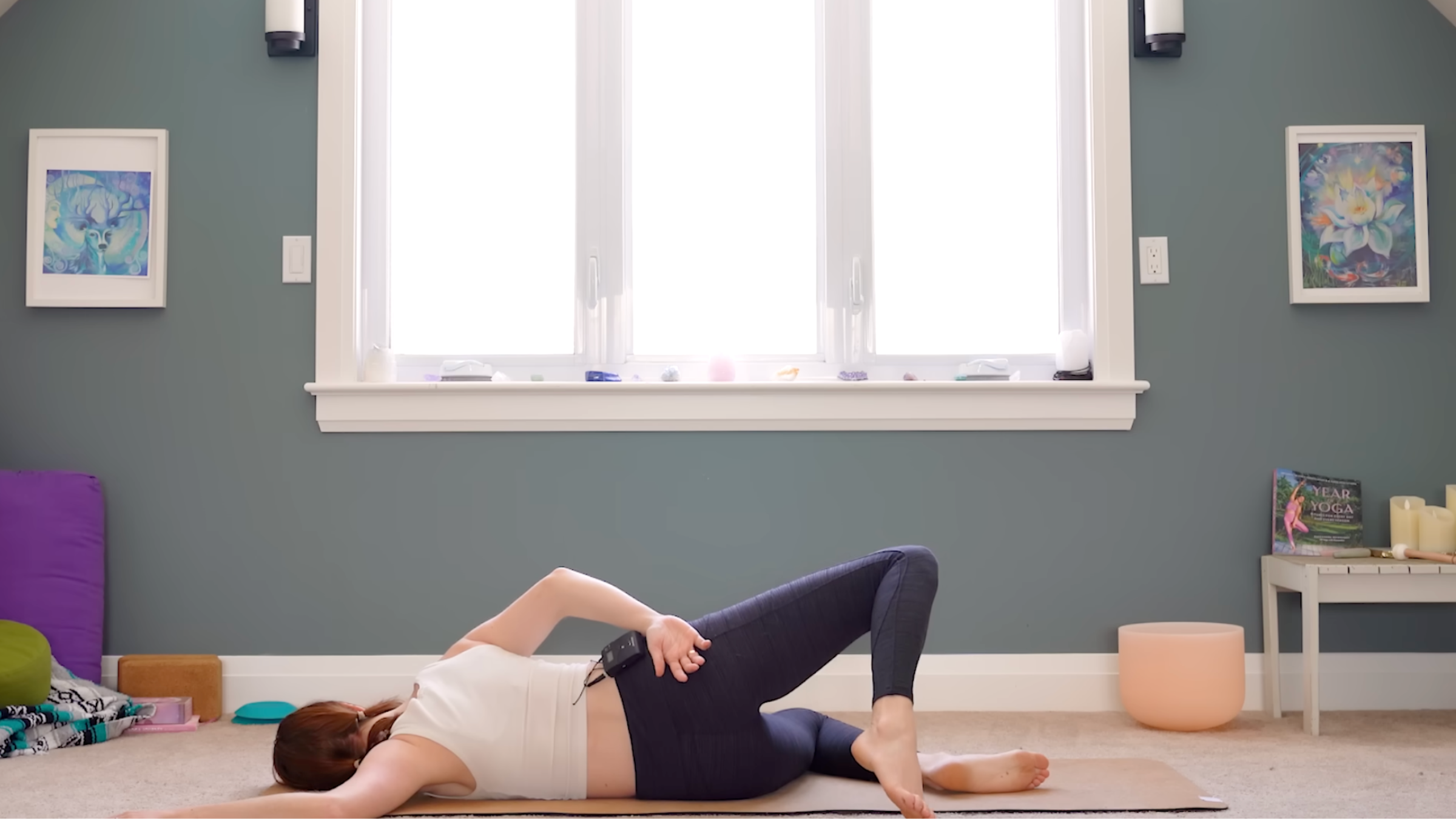 woman in white tank and black tights in side-lying shoulder stretch pose on left side with knees bent and left arm in 90-degree angle on floor, right leg bent and lifted, right arm on right hip, on tan yoga mat in carpeted room with windows and yoga accessories in the background