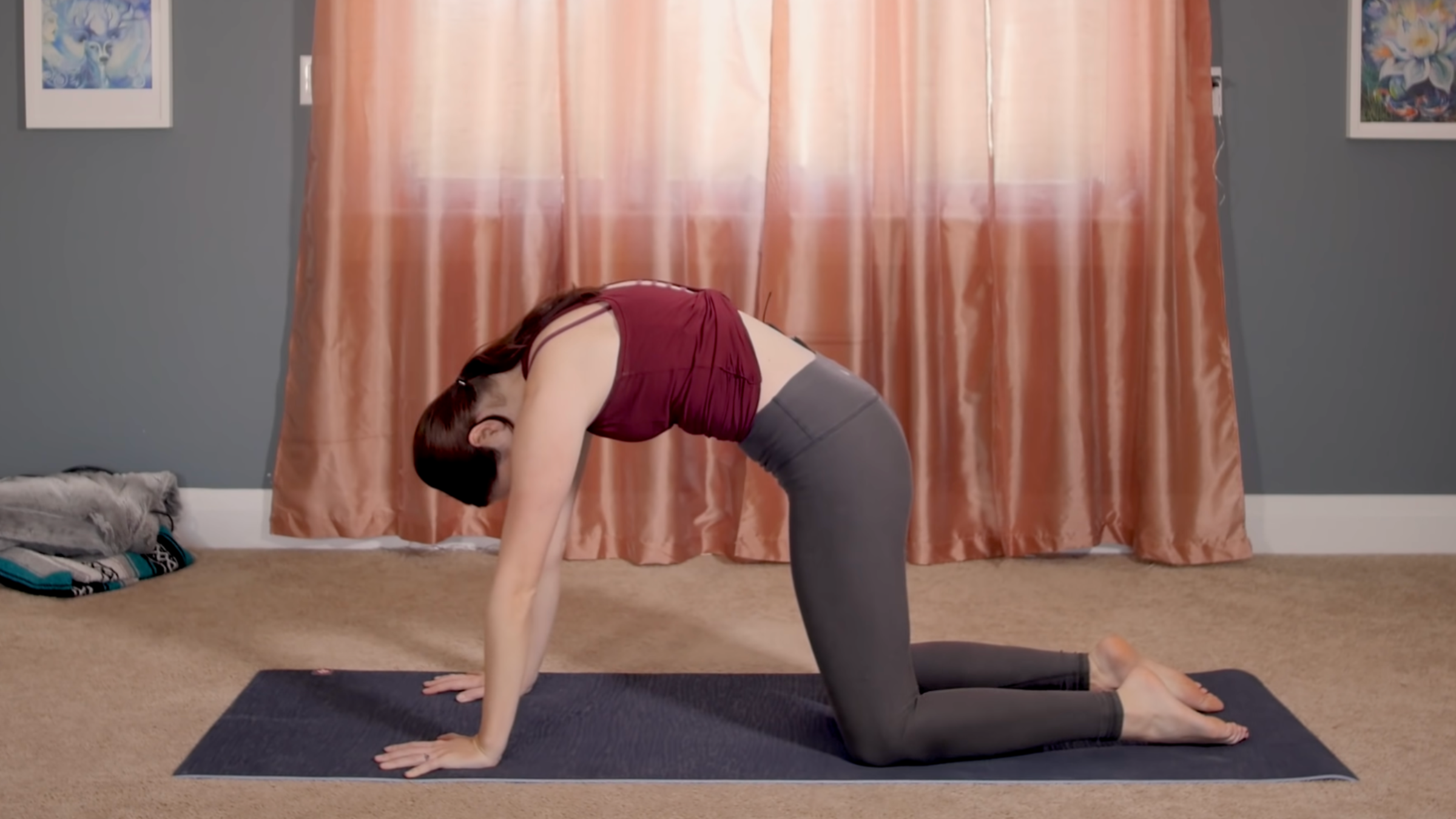 woman in yoga clothes in cat pose on yoga mat in home in front of pink curtains closed in front of window