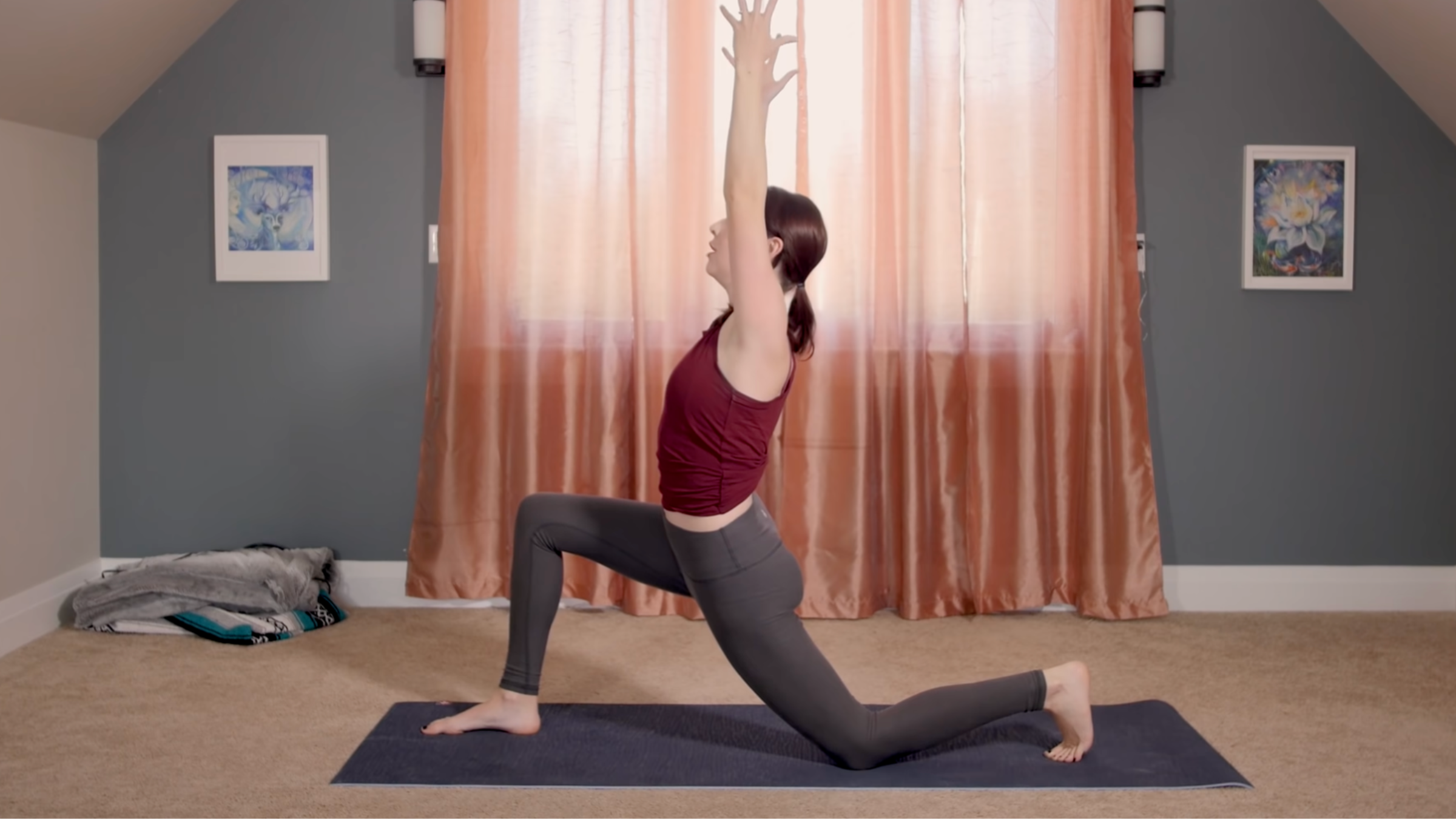 woman in low lunge pose with arms extended overhead and right leg forward on mat in home in front of pink curtains closed in front of window