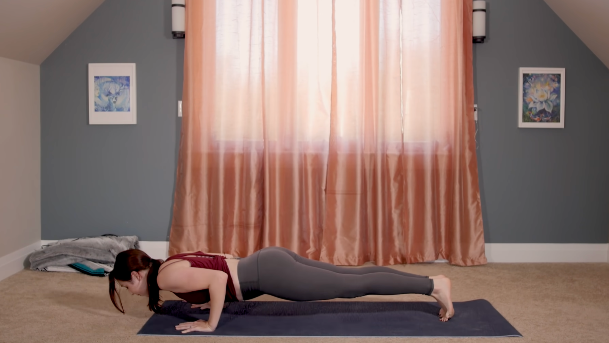 woman in yoga clothes in Chaturanga Dandasana pose, in a plank lowered halfway to the ground, on yoga mat in home in front of pink curtains closed in front of window