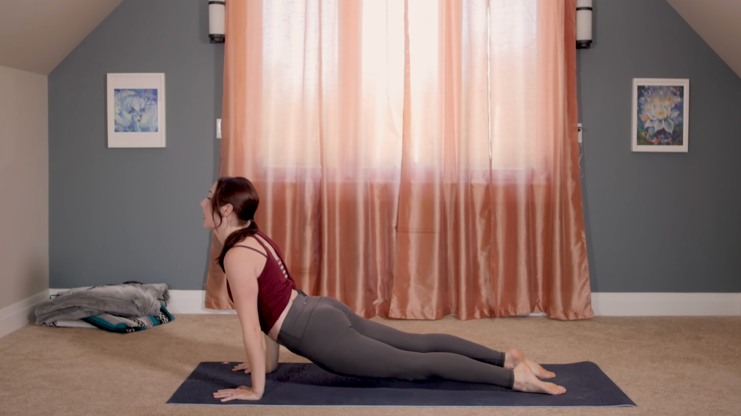 woman in yoga clothes in upward-facing dog pose on yoga mat in home in front of pink curtains closed in front of window
