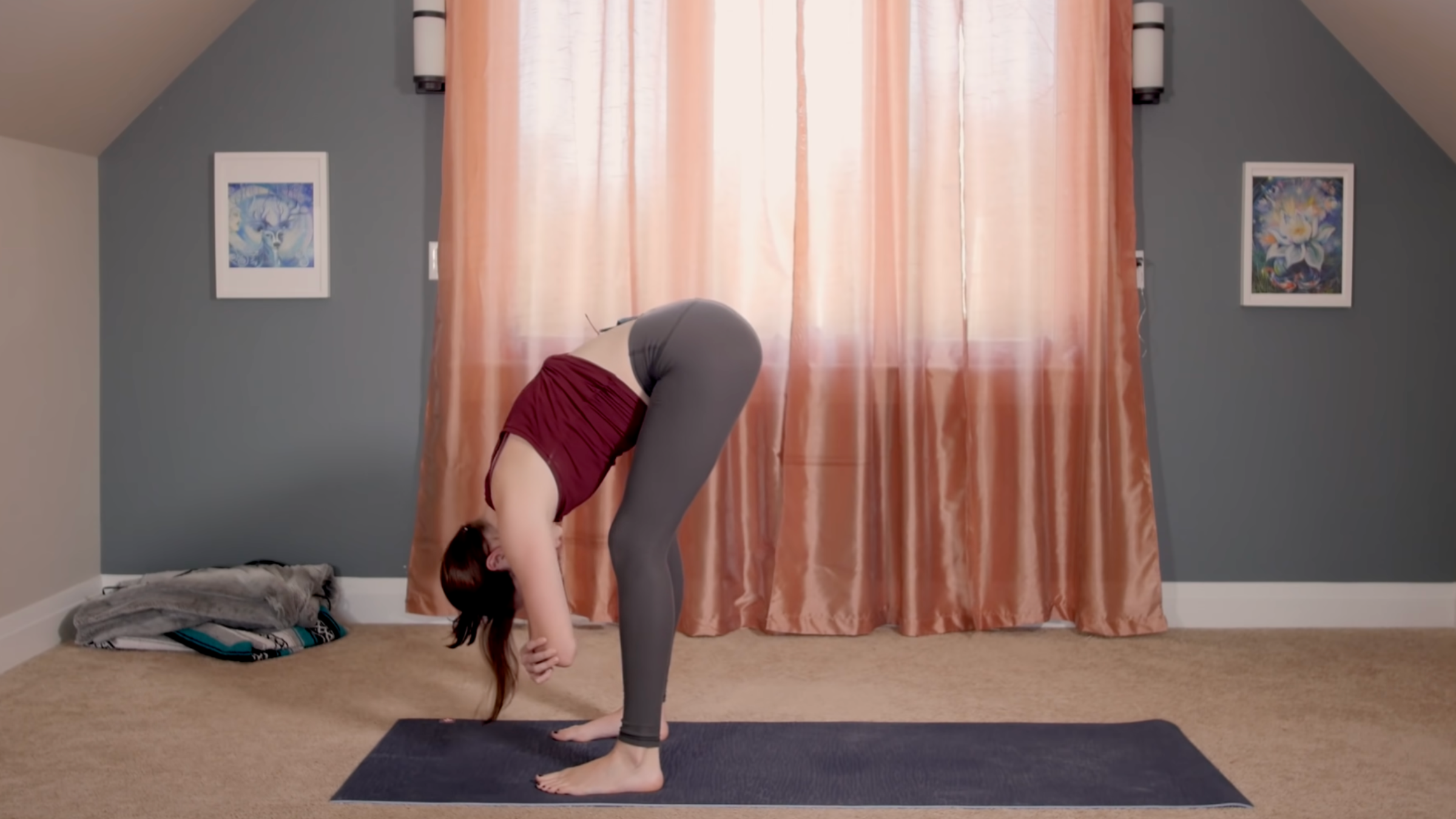 woman in yoga clothes in standing forward bend pose on yoga mat in home in front of closed curtains