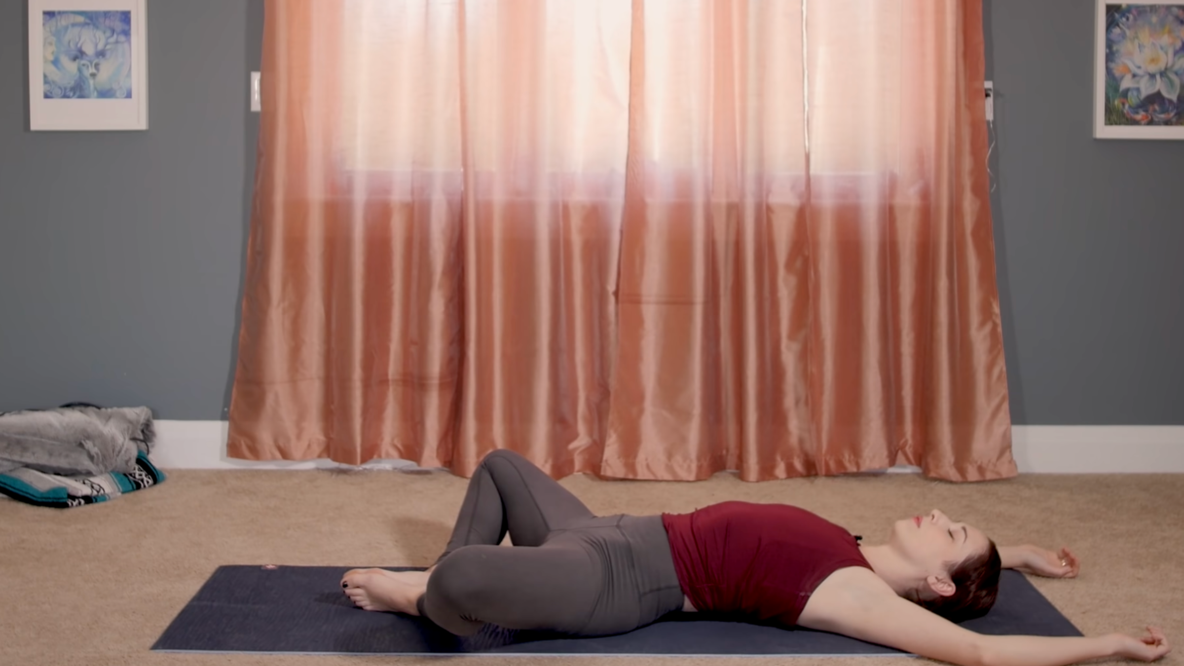 woman wearing clothes in reclined bound angle pose on mat in home in front of closed curtains