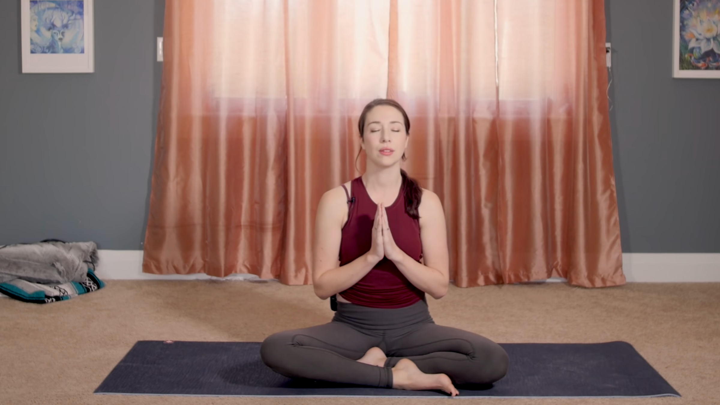 woman in seated meditation pose on yoga mat with legs crossed and hands in prayer at chest, in home in front of closed curtains