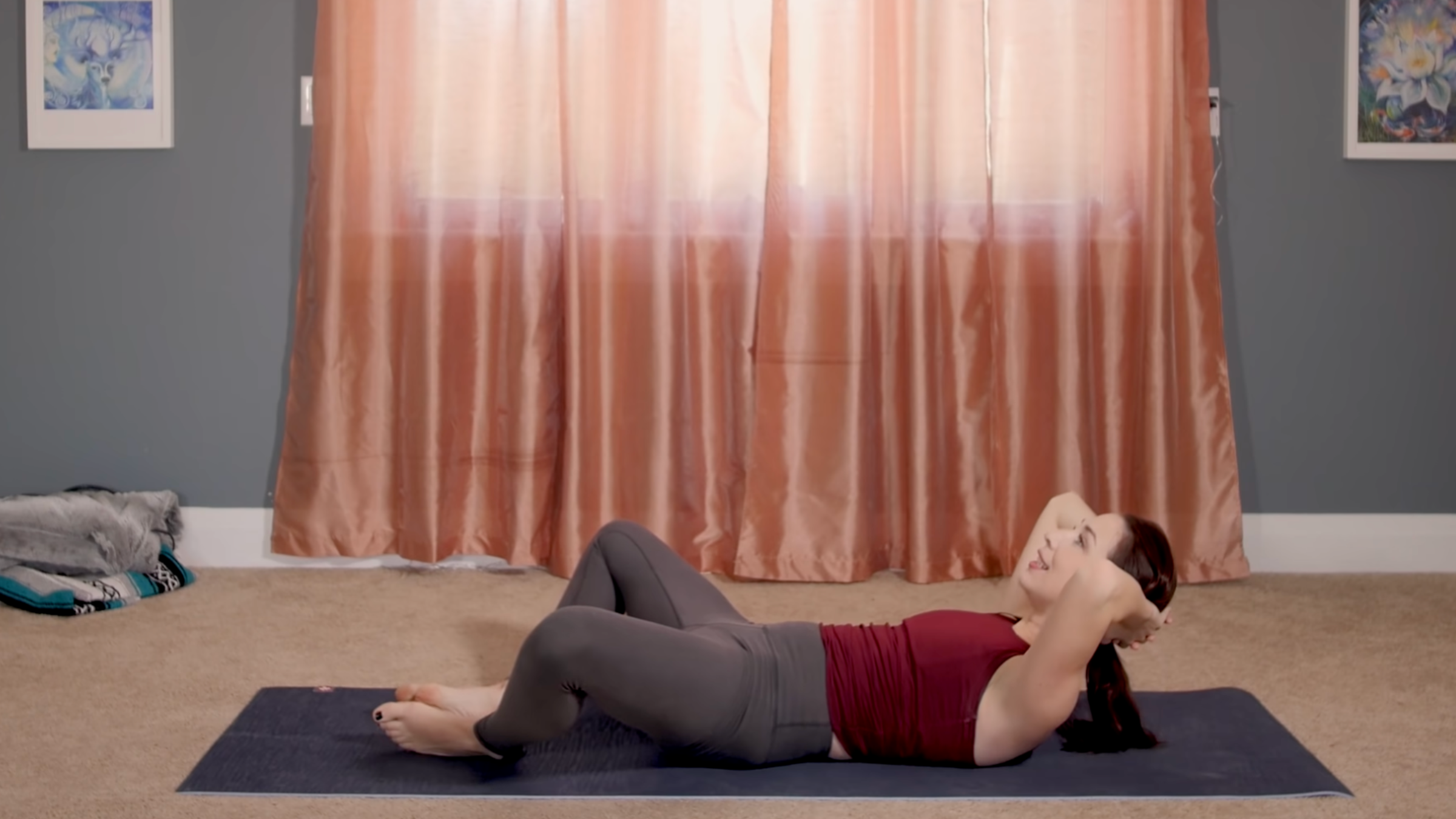 woman wearing yoga clothes in reclined crunch position with legs in cobbler pose on yoga mat in home in front of closed curtains