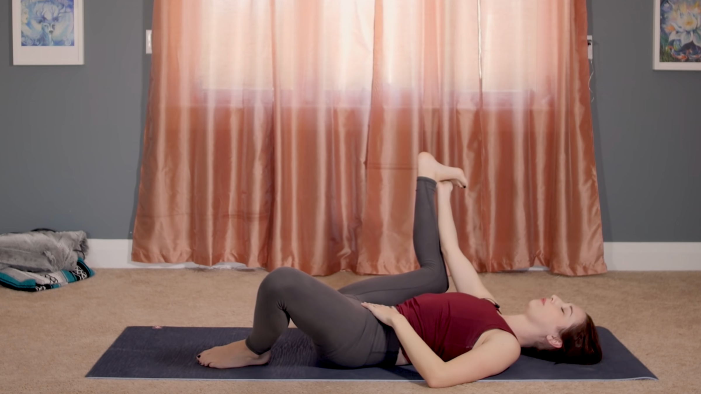 woman in yoga clothes in half happy baby pose on yoga mat in home in front of closed curtains