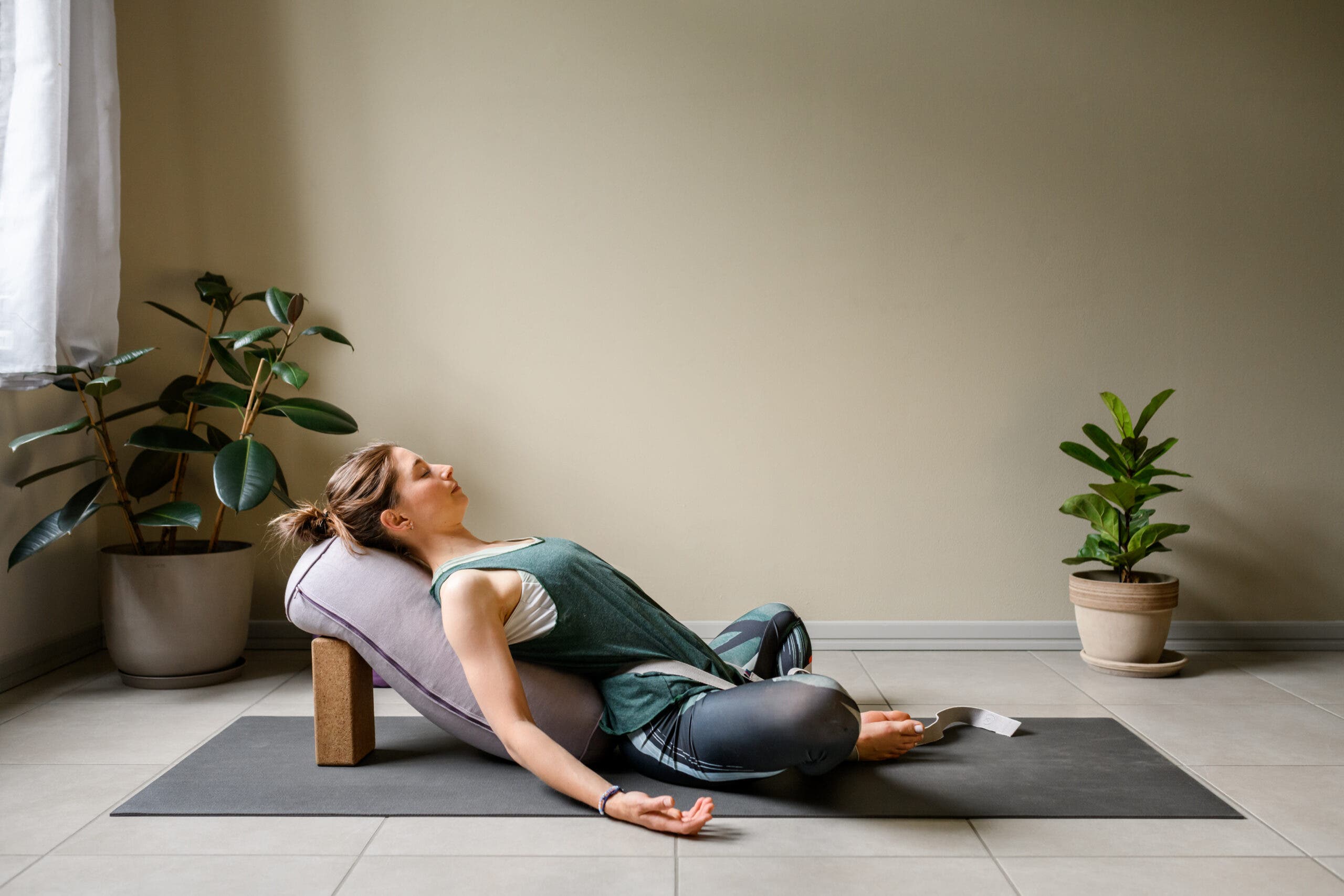 Woman practicing reclining bound angle pose with a bolster underneath her back. She's in a dimly lit room with houseplants in the background.