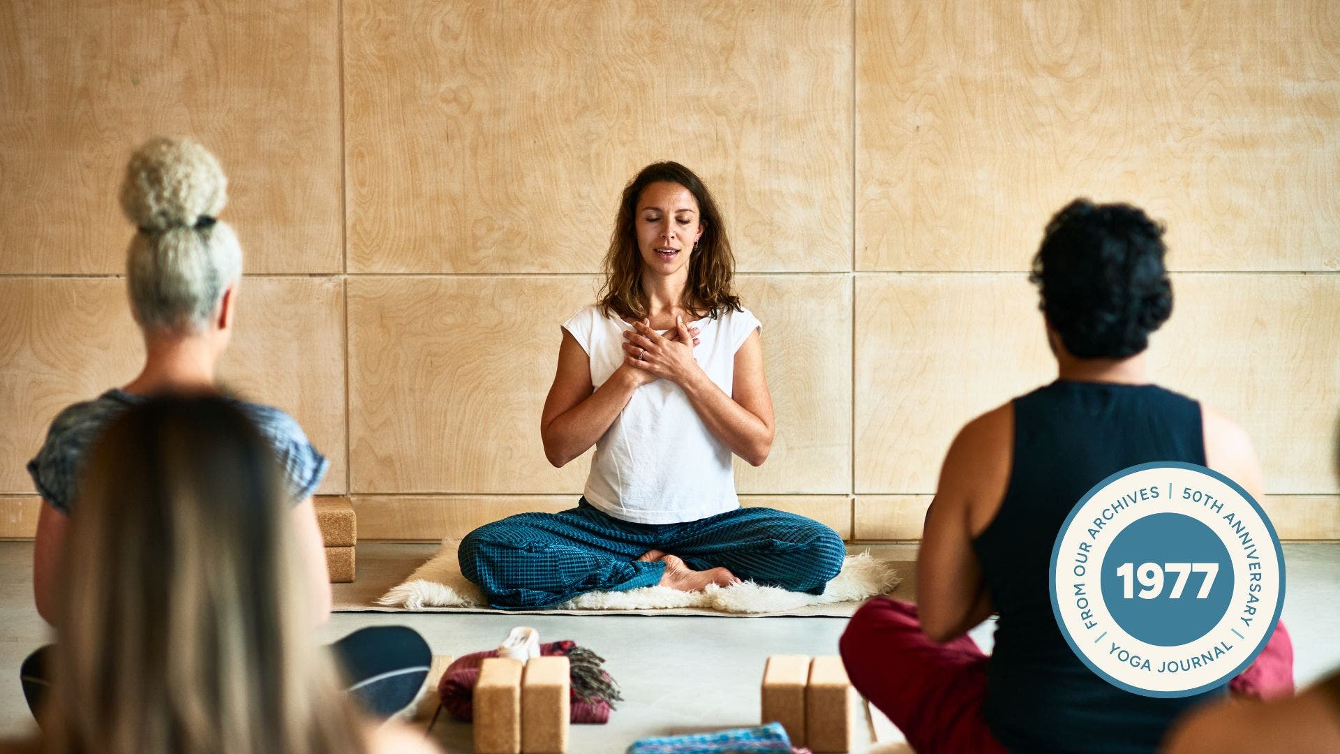 Image of yoga teacher sitting cross-legged with her eyes closed and hands over her chest with two yoga students looking at her.