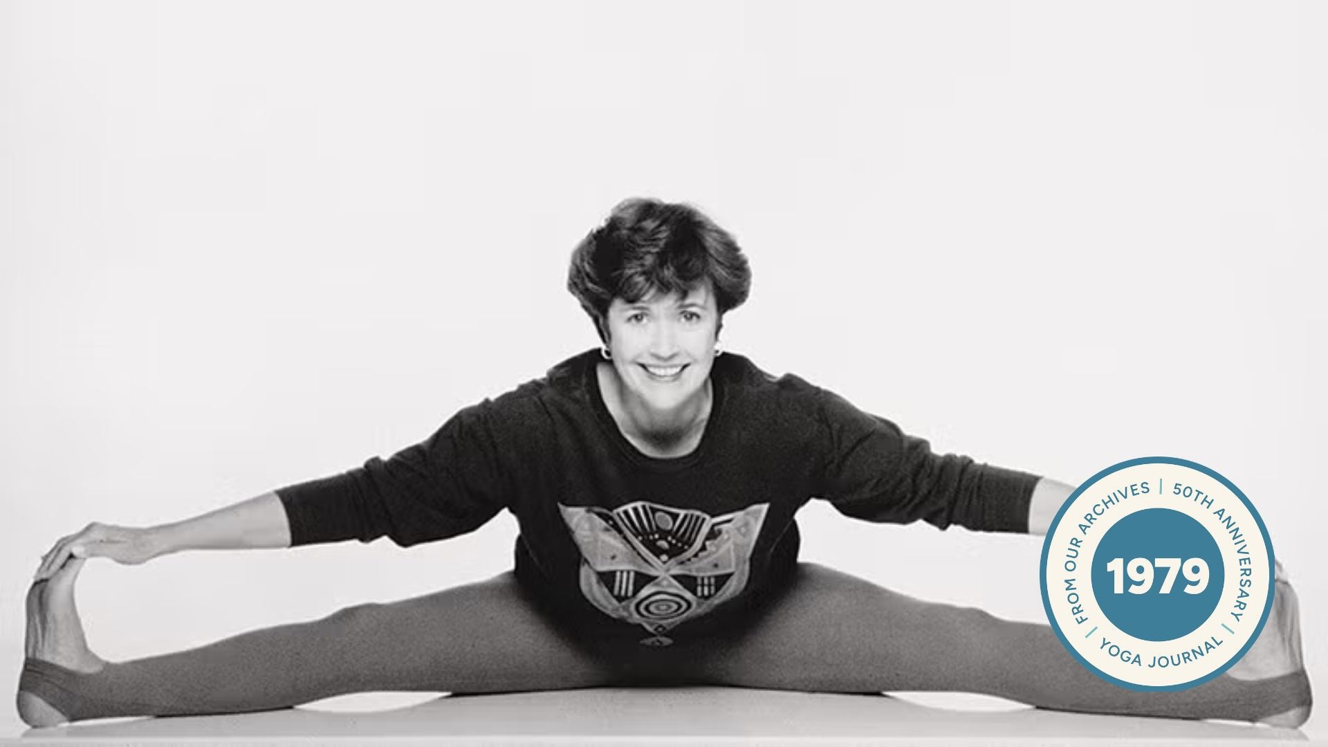 Black and white image of Lilias Folan smiling in Wide-Legged Seated Forward Bend.