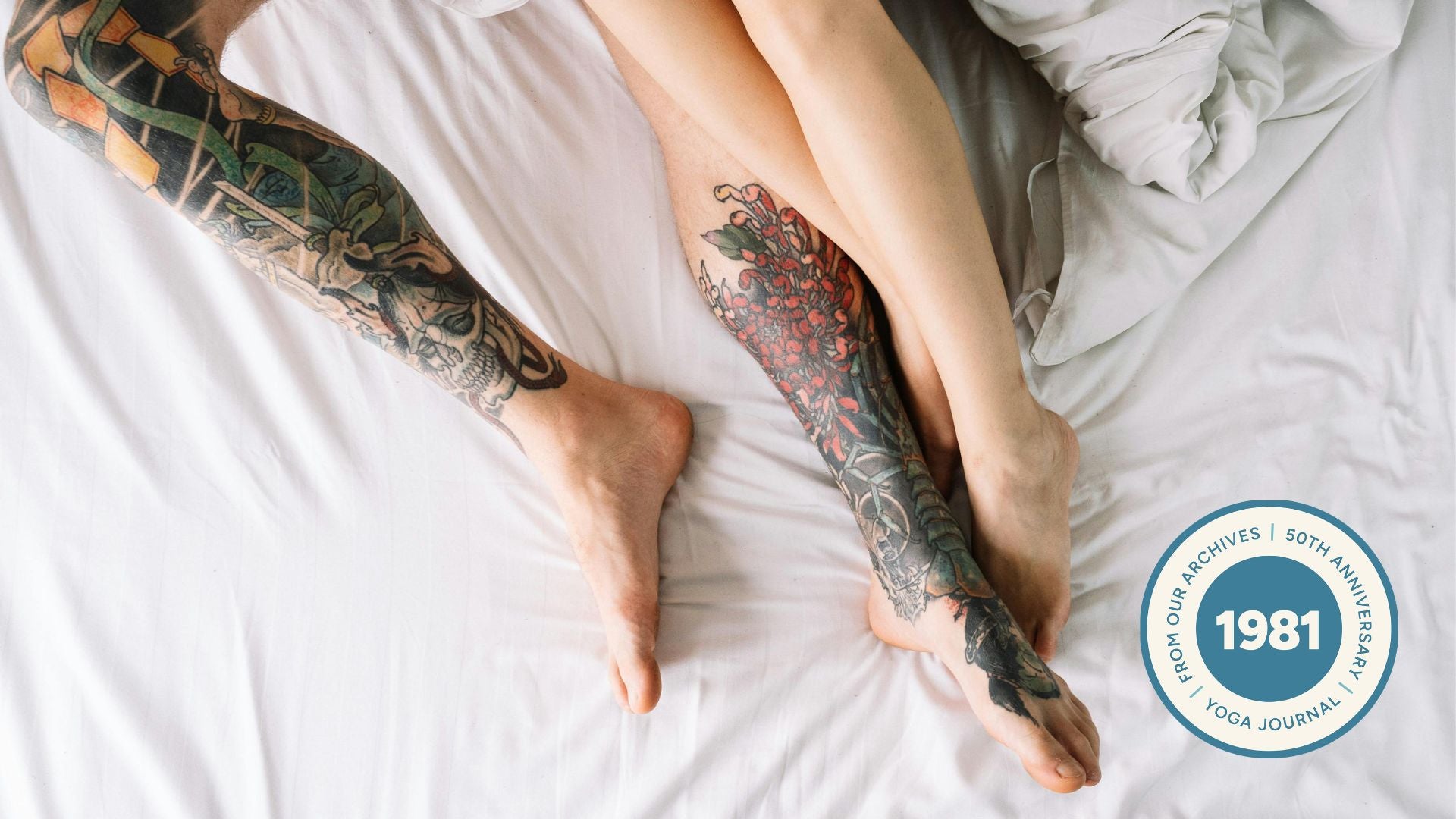 Image of couple legs intertwined in bed. One of them has colorful tattoos in their legs.