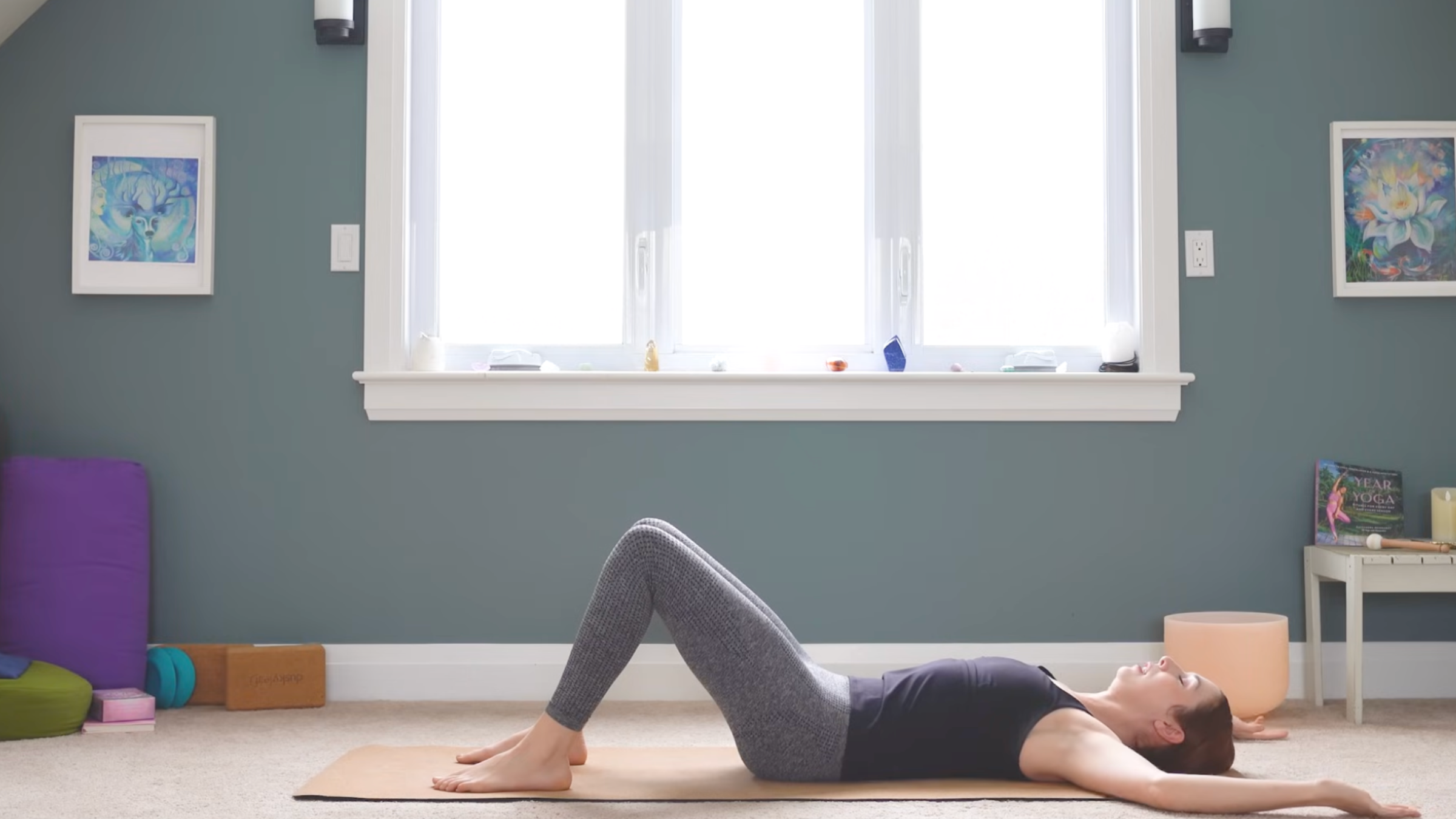 woman in yoga clothes lies on back with knees bent and feet on the yoga mat in home in front of windows