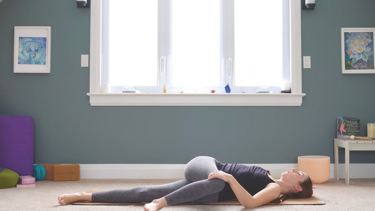 woman in reclined twist on yoga mat, lying down right left leg extended and right knee bent and crossed over to floor on left side of mat, with left hand holding down right knee