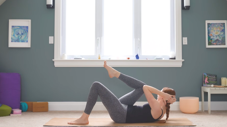woman in crunch twist with fingers interlaced behind head and lifting up head and shoulders, left foot on yoga mat, and left elbow touching right knee
