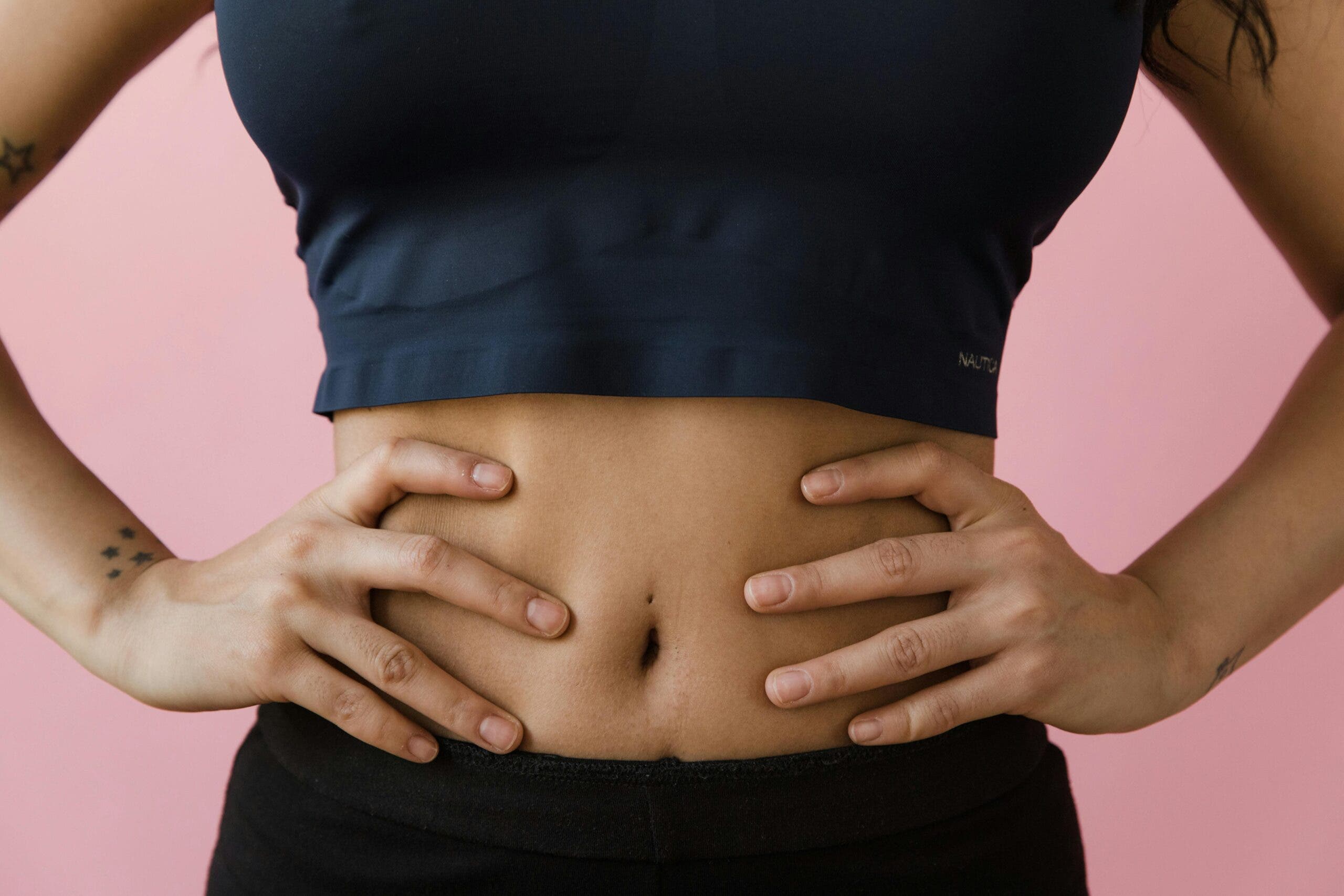 Close-up of woman's bare stomach. She is wearing workout clothes and her hands are on her hips. Standing in front of a bubblegum pink background.