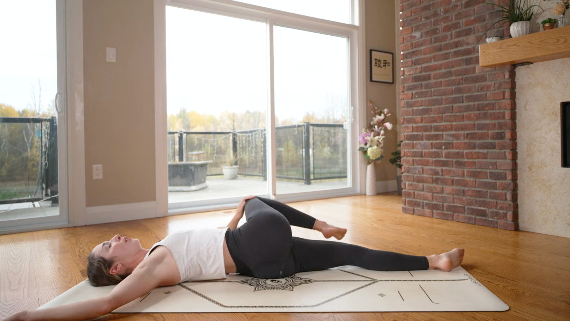 Yoga teacher lying on her back on a mat in a reclined twist with her right leg bent and drawn toward the left