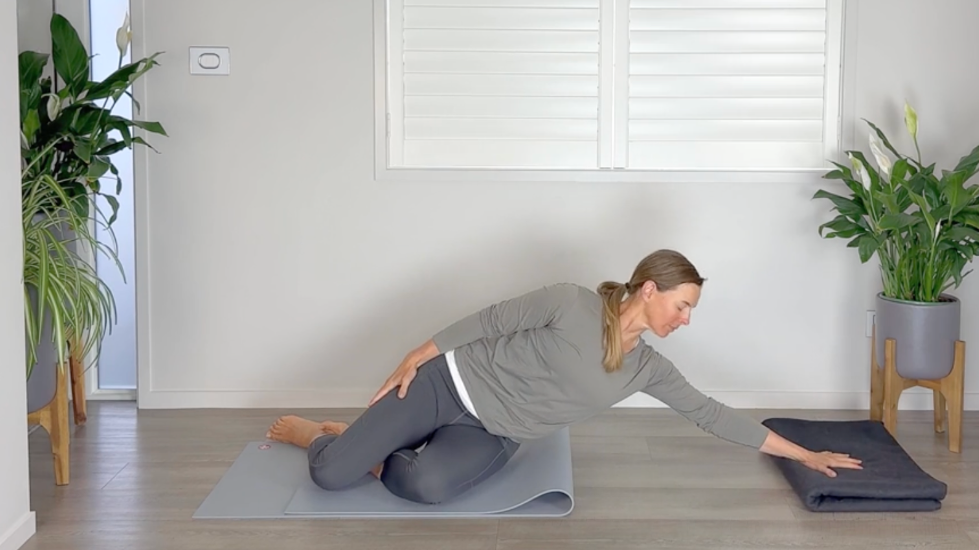 A woman sits on a mat with her left hand on a folded yoga blanket as she turns away in a side stretch