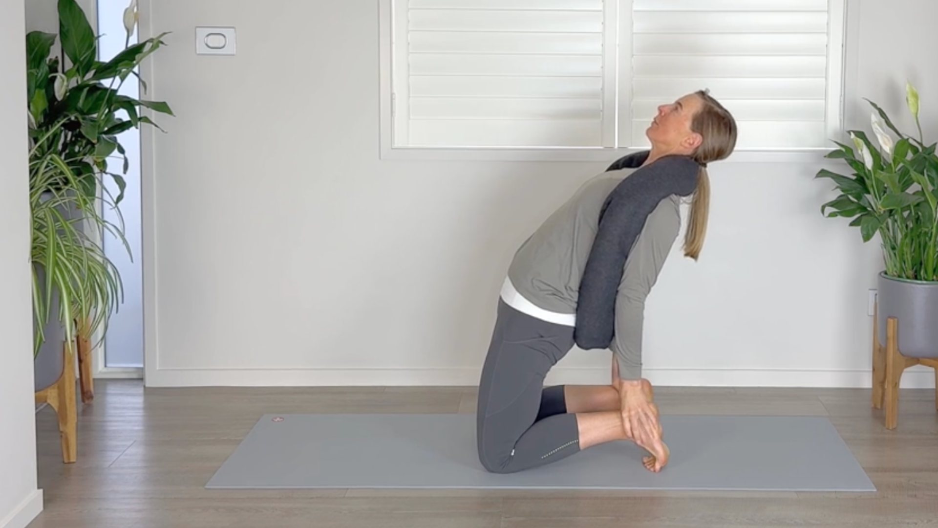Woman kneels on a mat in camel pose with a yoga blanket wrapped around the back of her neck for support