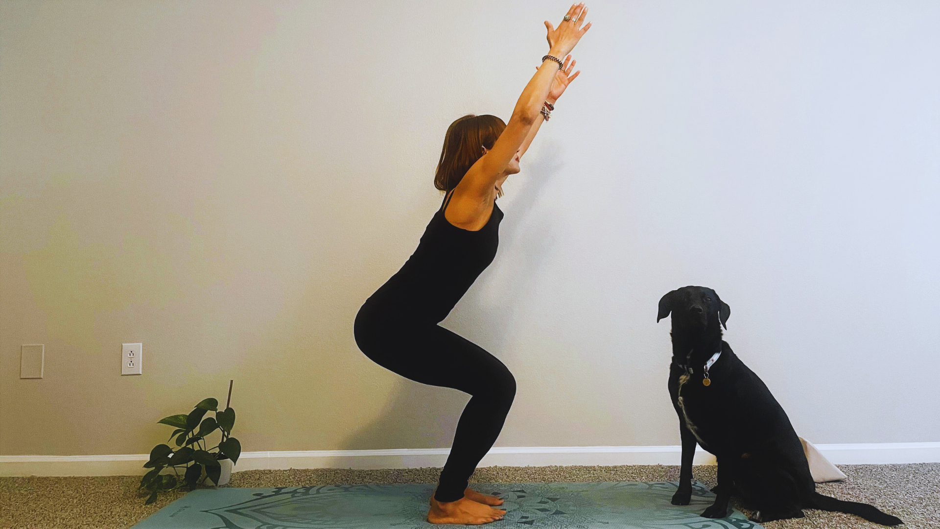 Woman standing alongside her dog while sitting back in Chair Pose