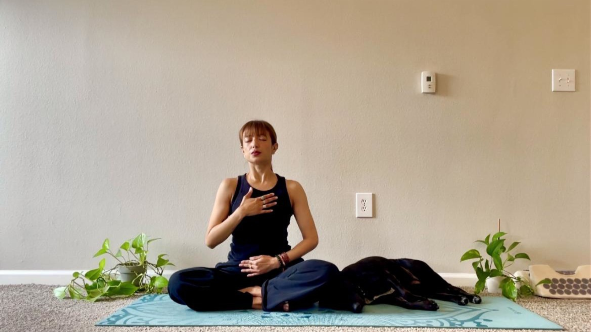 Yoga teacher seated on a mat with one hand on her belly and the other on her chest practicing box breathing (sama vritti).