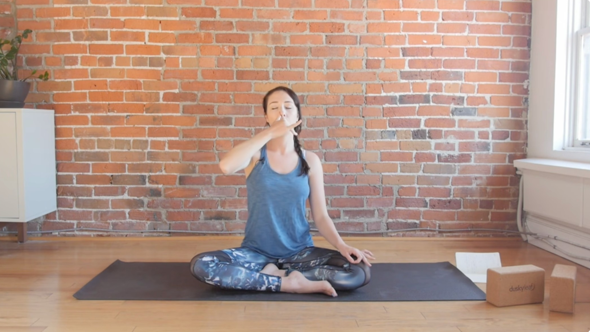 Woman sitting cross-legged on a yoga mat practicing alternate nostril breathing or nadi shodhana by closing off one nostril while breathing through the other